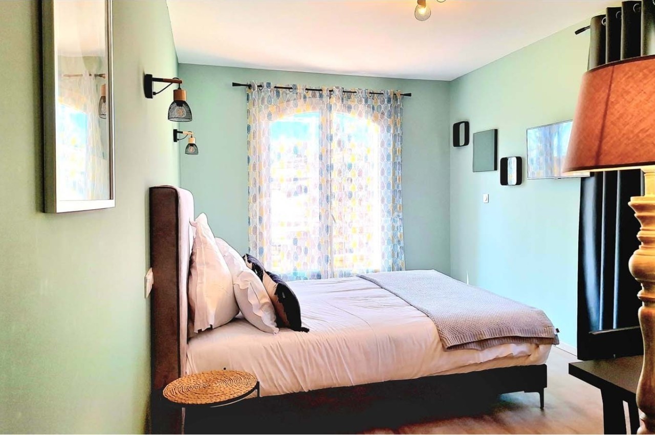 Chambre lumineuse avec lit double, murs verts, rideaux à motifs, lampe de chevet et décoration murale moderne.