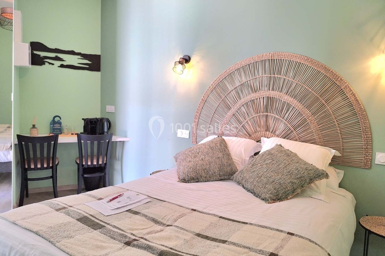 Chambre lumineuse avec un lit double, tête de lit en rotin, bureau avec chaises et mur peint en vert clair.