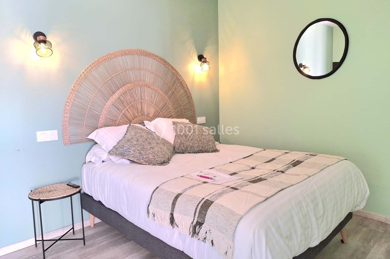 Lit double avec tête de lit en rotin, plaid beige, coussins assortis, table d'appoint et mur vert pastel.