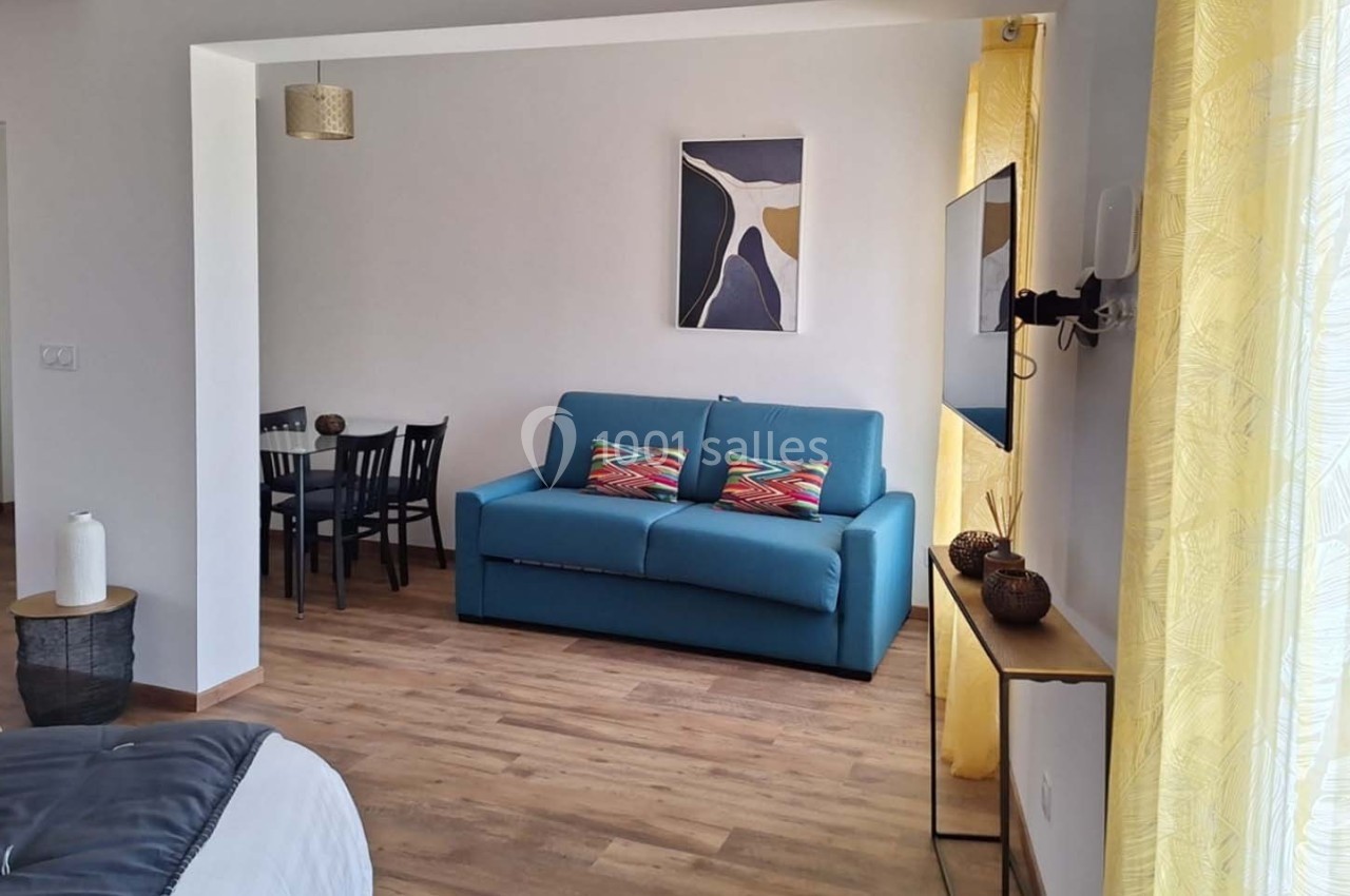 Salon lumineux avec canapé bleu, table à manger noire, parquet en bois et rideaux jaunes laissant passer la lumière.