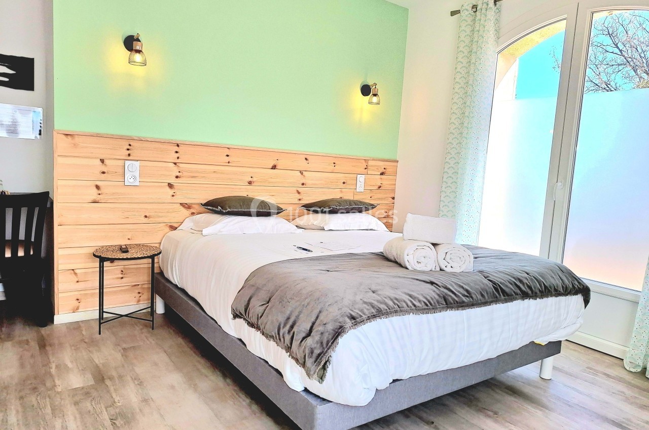 Chambre lumineuse avec lit double, tête de lit en bois, murs verts et blancs, et grande fenêtre avec rideaux clairs.