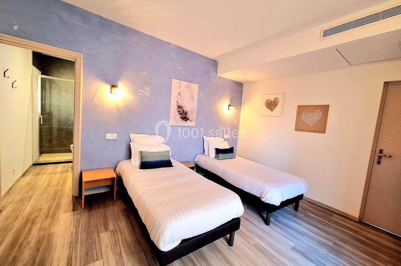 Chambre lumineuse avec deux lits simples, parquet, mur bleu, décorations murales et salle de bain attenante.