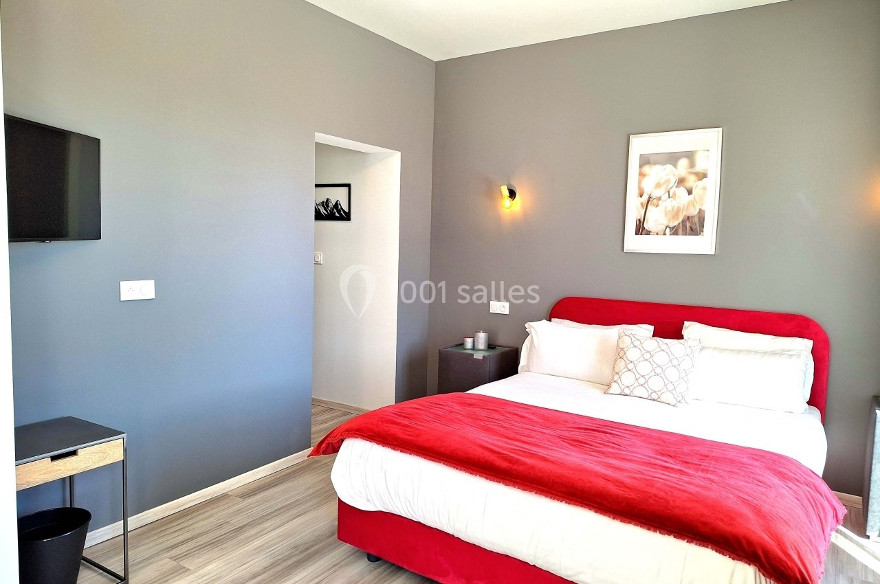 Chambre moderne avec lit double, mur gris, linge rouge et blanc, télévision murale et éclairage minimaliste.