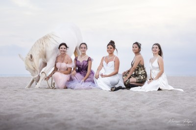 Deux femmes en costumes ornés posent à côté d'un cheval blanc dans un jardin ensoleillé.