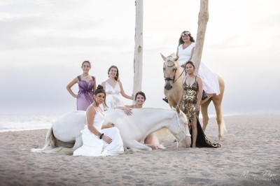 Deux femmes en costumes ornés posent à côté d'un cheval blanc dans un jardin ensoleillé.