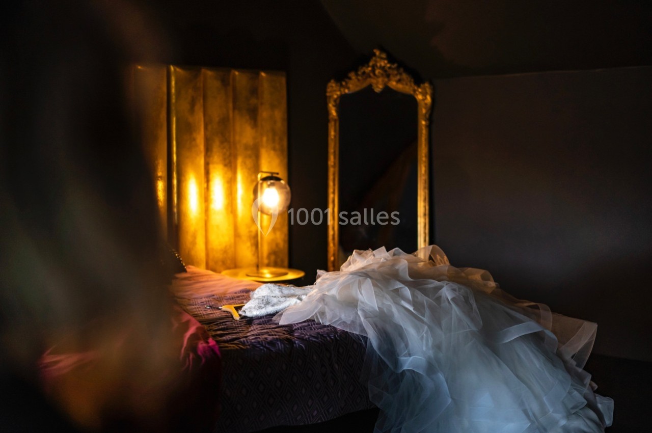 Robe blanche en tulle posée sur un lit, près d'un miroir doré et d'une lampe allumée dans une pièce sombre.