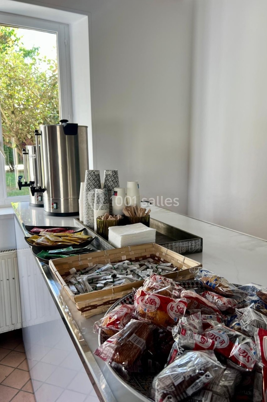 Table avec boissons chaudes, gobelets, sucre, biscuits et condiments, près d'une fenêtre donnant sur un jardin.