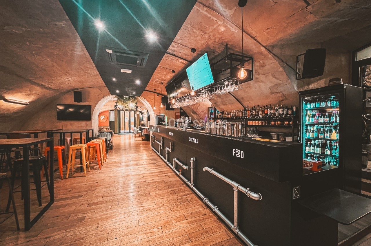 Intérieur d'un bar moderne avec comptoir noir, tabourets colorés, étagères de bouteilles et éclairage chaleureux.