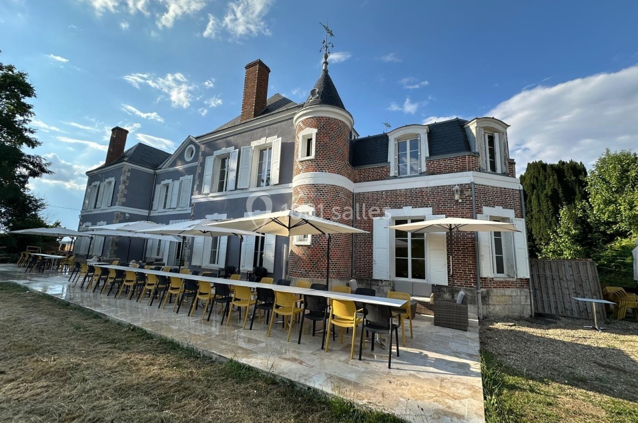 Façade d'une grande maison en briques et pierre avec terrasse aménagée de tables, chaises et parasols.