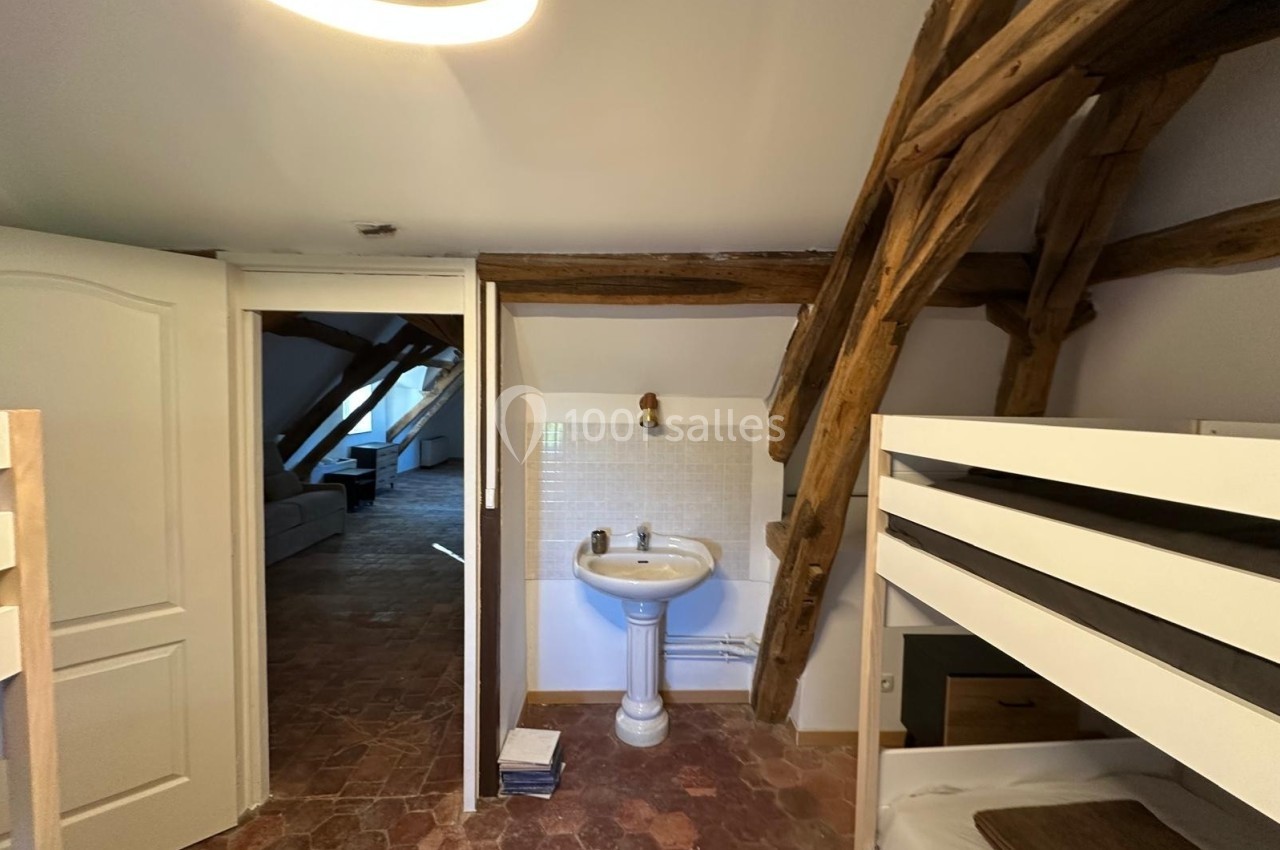 Lavabo blanc avec miroir dans une pièce mansardée, poutres apparentes et lits superposés visibles au premier plan.