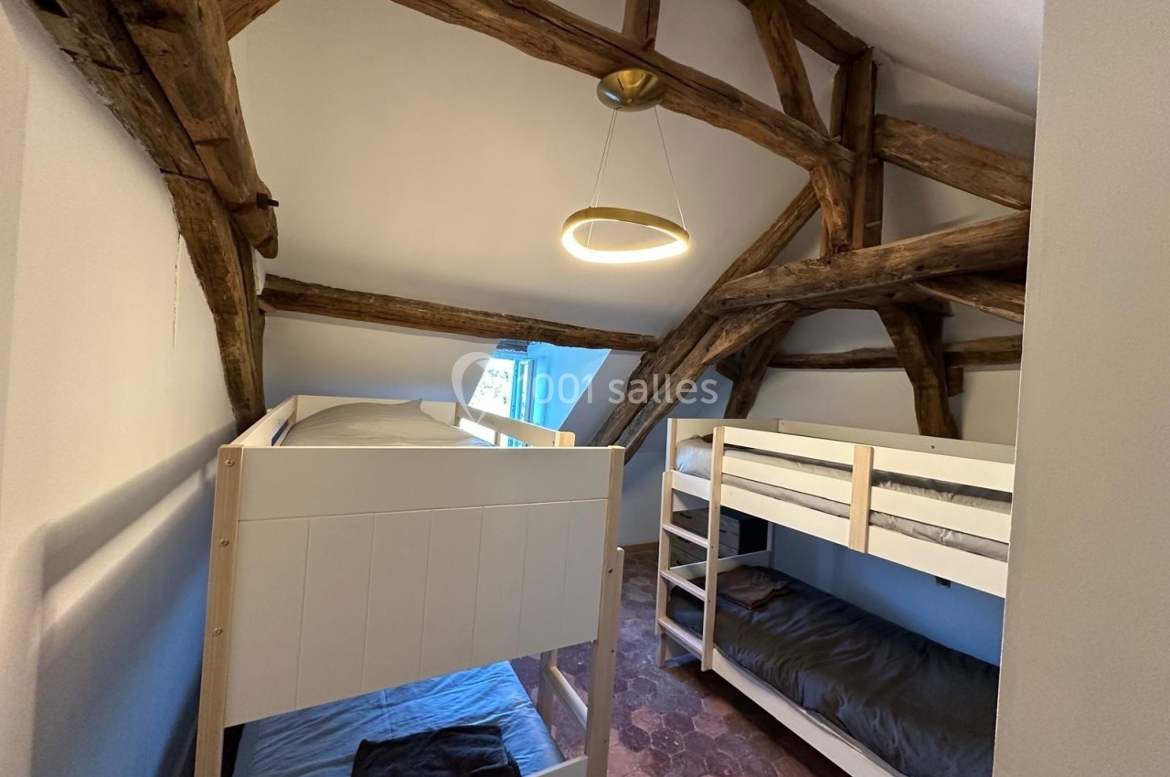Chambre mansardée avec poutres apparentes, équipée de deux lits superposés et éclairée par une fenêtre et un luminaire…