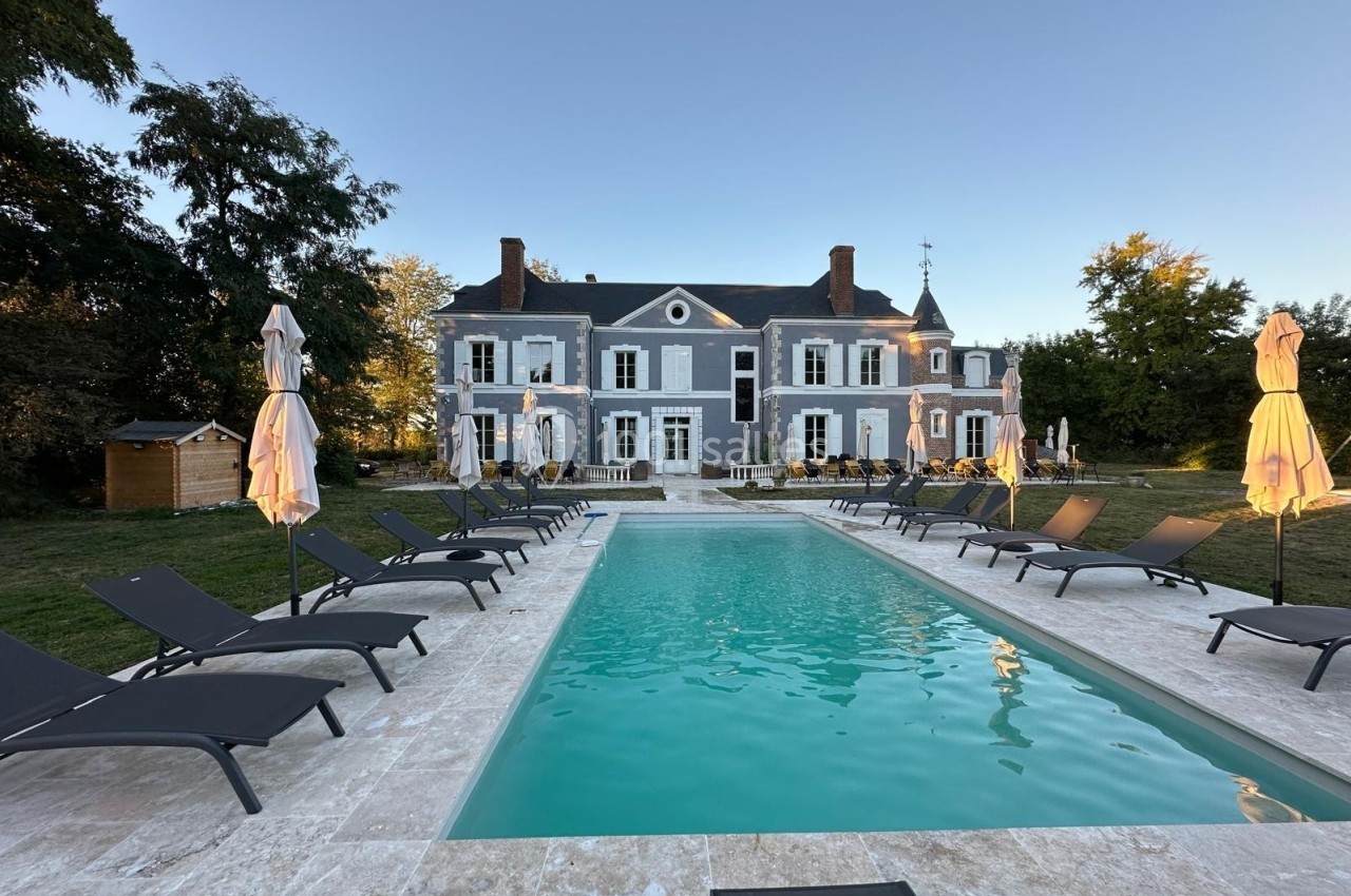 Piscine extérieure entourée de chaises longues et parasols, devant une grande maison de style classique.