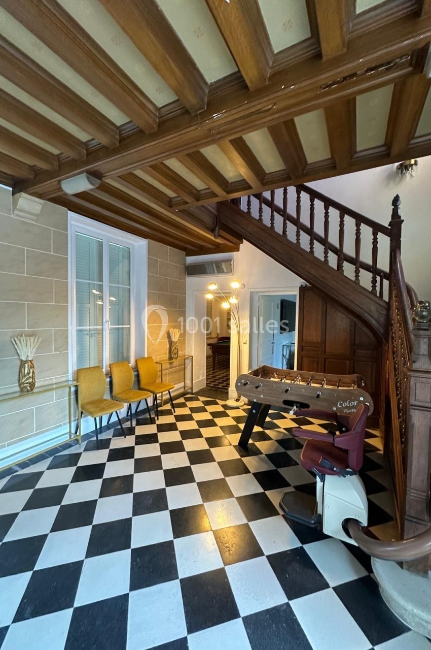 Entrée avec escalier en bois, sol en damier noir et blanc, baby-foot, fauteuil et chaises jaunes près d'une table.