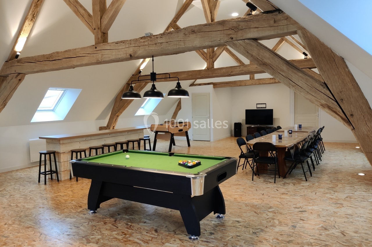 Salle lumineuse sous combles avec billard, baby-foot, grande table de réunion et poutres apparentes.