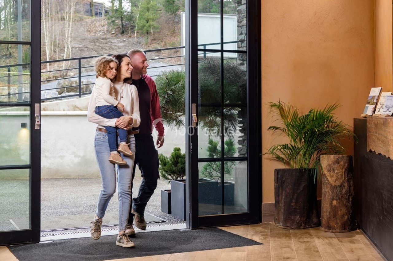 Une famille entre dans un bâtiment moderne avec de grandes baies vitrées, entouré de plantes et de bois naturel.