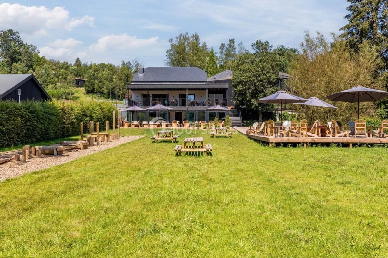 Espace extérieur avec tables de pique-nique, terrasse en bois, parasols et aire de jeux, entouré de verdure.