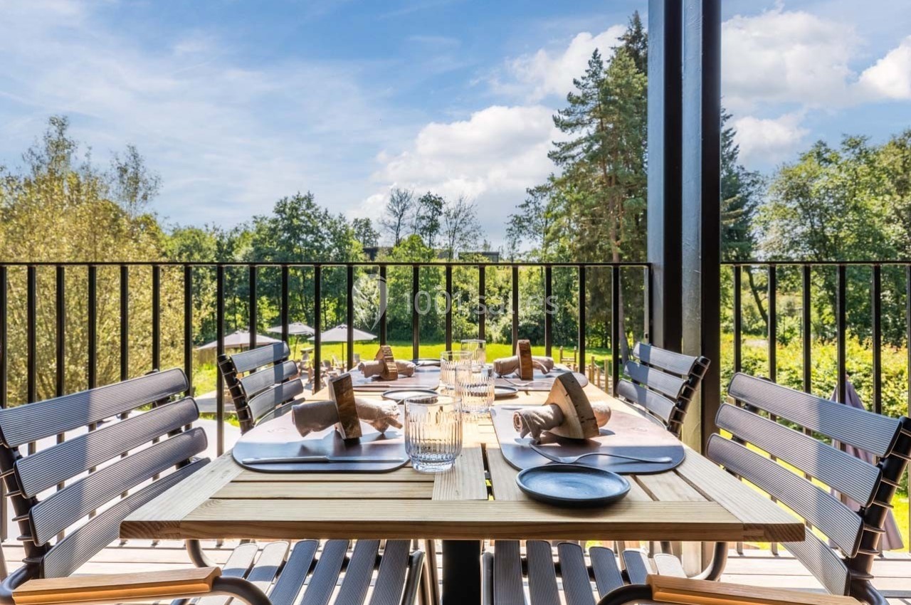 Table en bois dressée avec des assiettes, verres et serviettes sur une terrasse surplombant un jardin verdoyant.