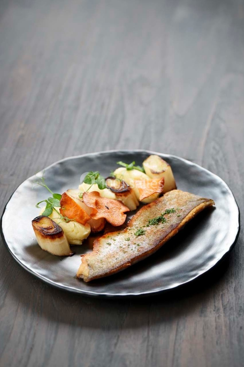 Filet de poisson grillé servi avec des légumes rôtis et des chips de légumes sur une assiette noire.