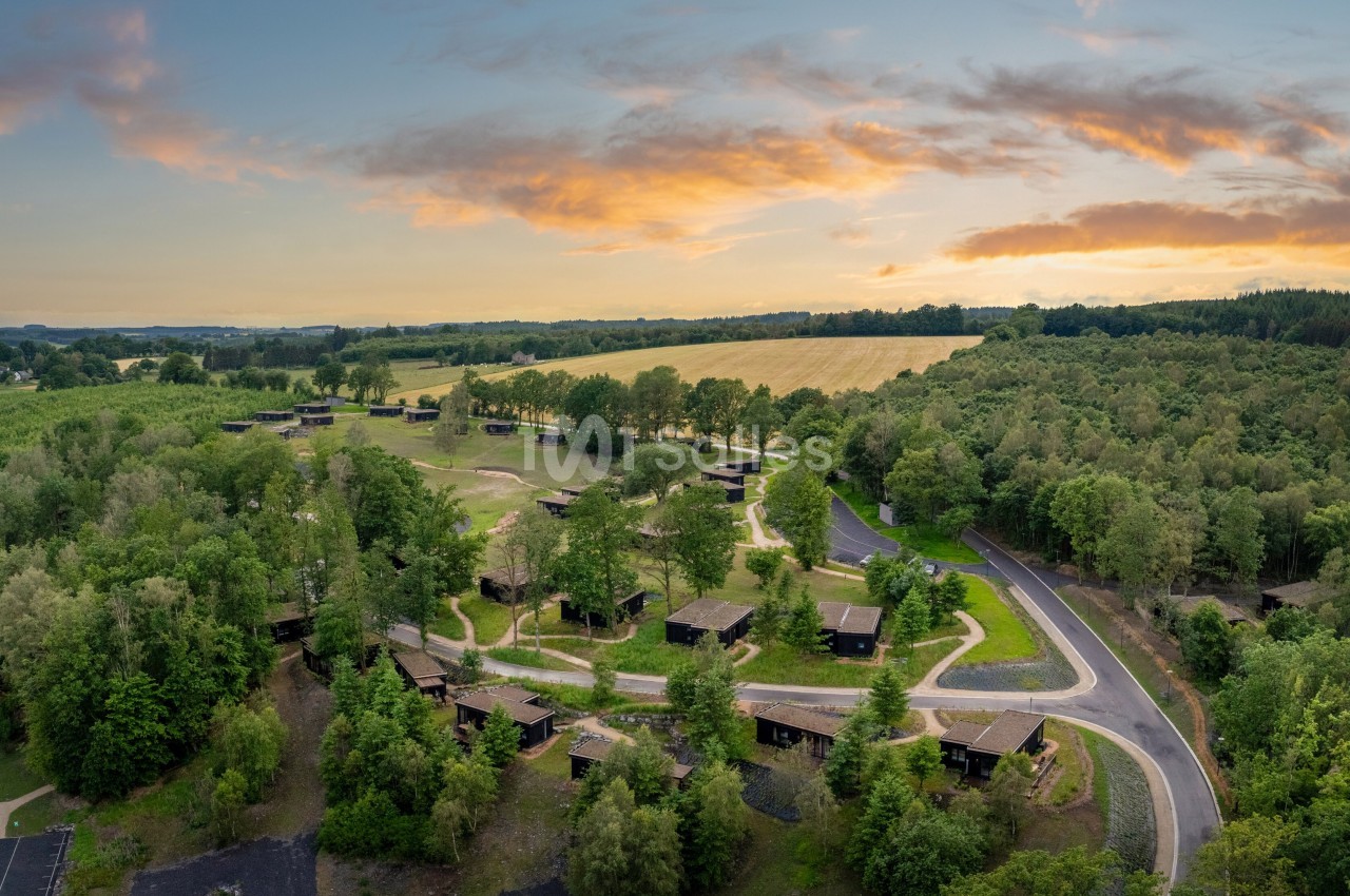 Vue aérienne d'un domaine de chalets en bois sombres dispersés dans une clairière boisée, sous un ciel de coucher de soleil orangé. Wait: must not start with "Vue de". The phrase starts with "Vue aérienne d'un domaine..." which is a forbidden start