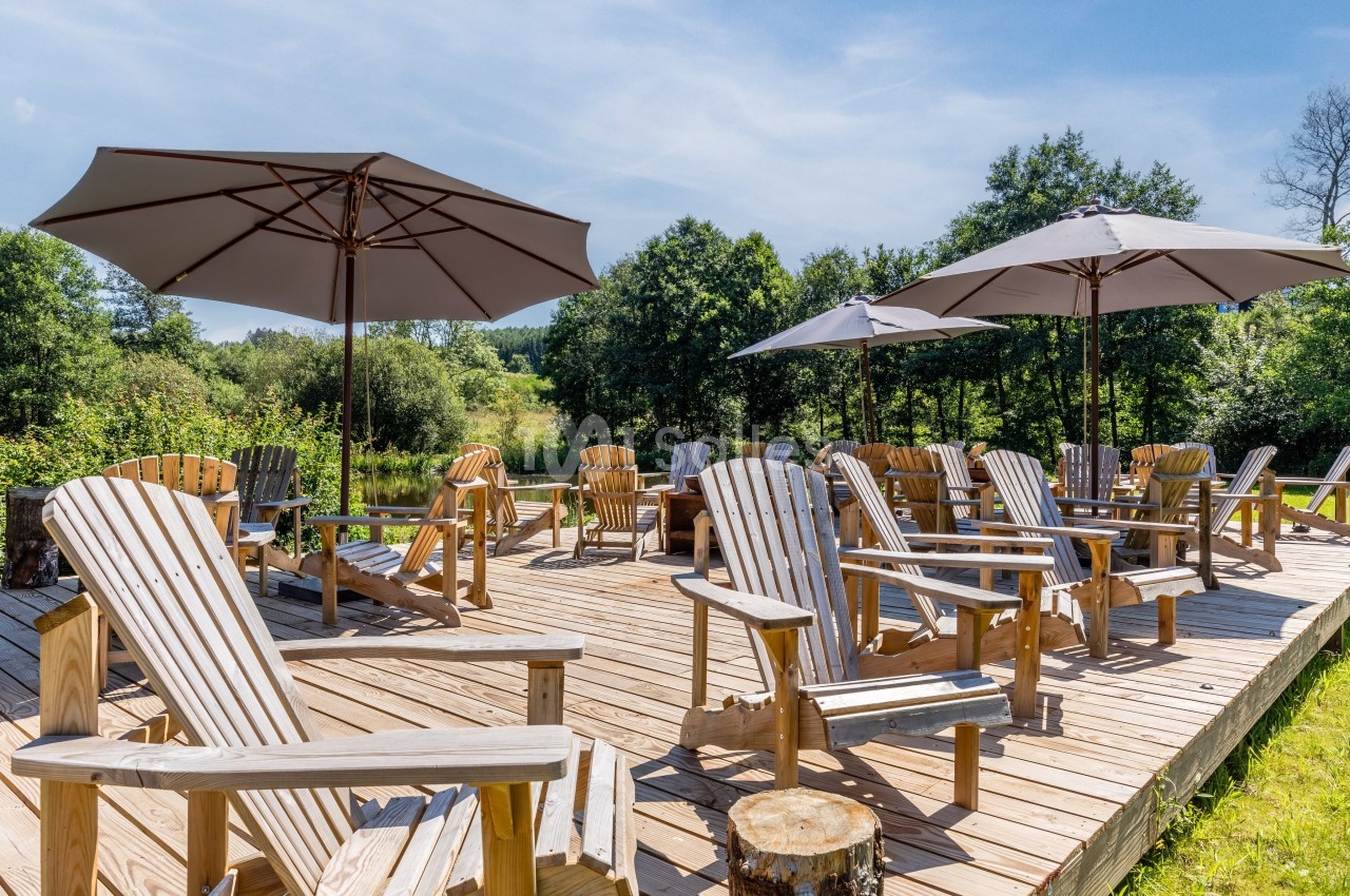 Terrasse en bois en bord d'eau avec des fauteuils Adirondack en bois clair disposés sous de grands parasols beige, entourée d'arbres.