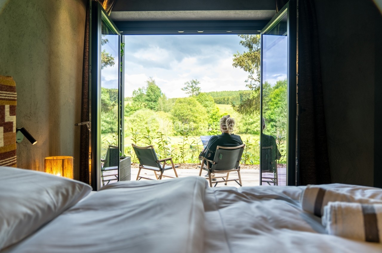 Vue depuis une chambre sur une terrasse avec une personne assise face à un paysage verdoyant sous un ciel nuageux.