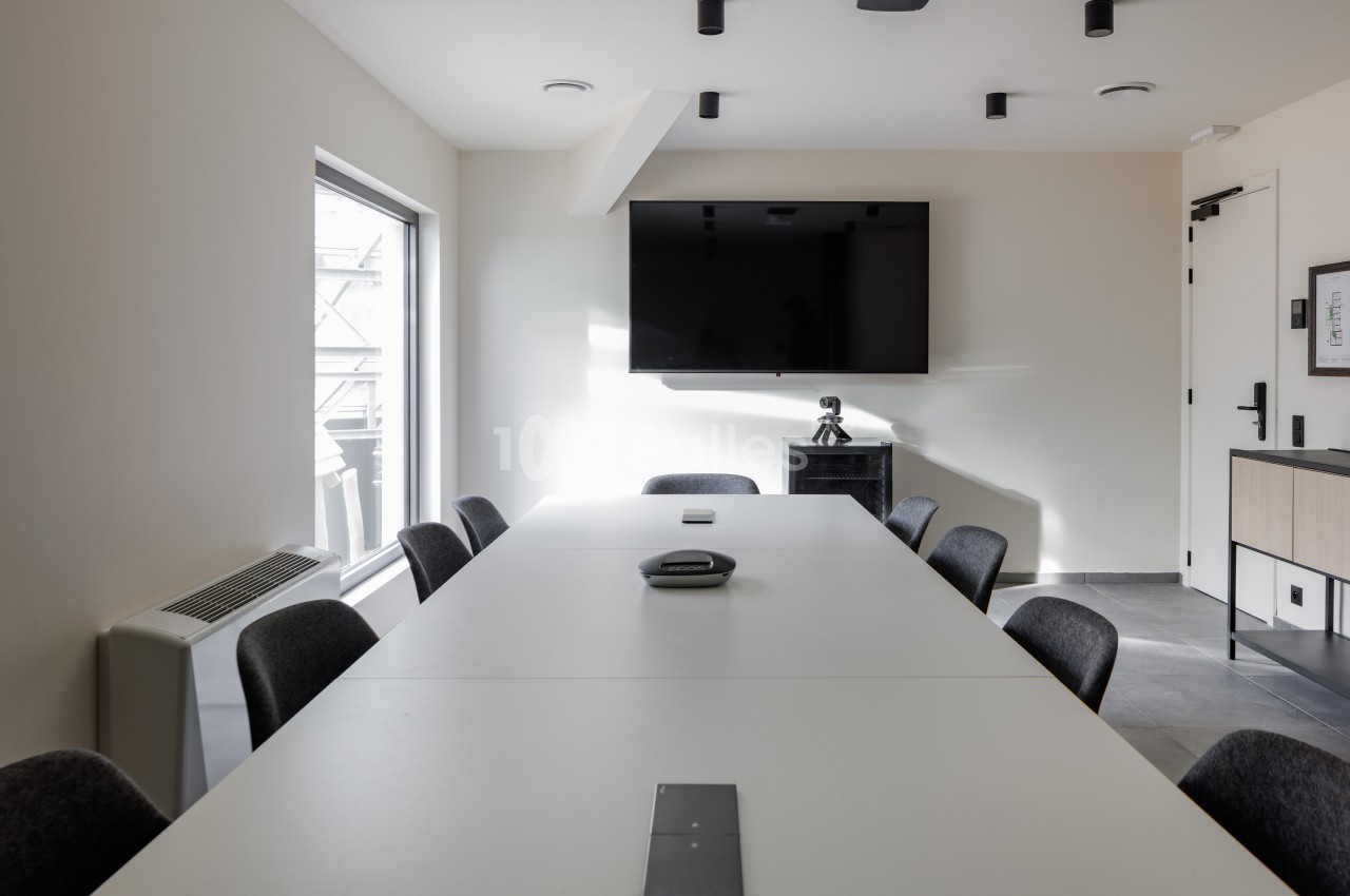 Salle de réunion lumineuse avec une grande table blanche, des chaises noires, un écran mural et une fenêtre.