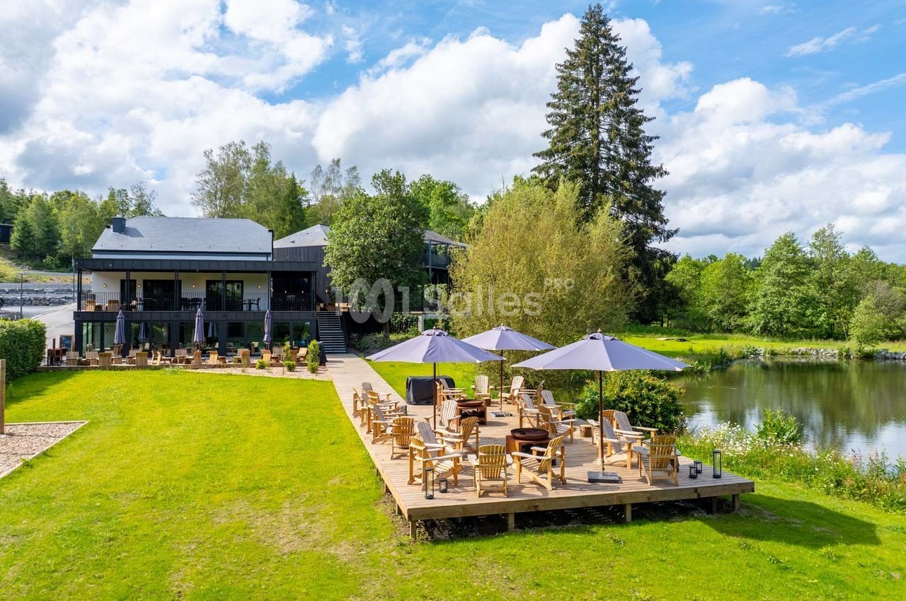 Terrasse en bois avec tables et parasols, bordant un étang entouré de verdure et d'arbres sous un ciel partiellement nuageux.