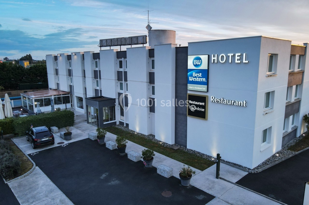 Façade d'un hôtel moderne avec enseignes lumineuses, parking et terrasse extérieure visibles au premier plan.