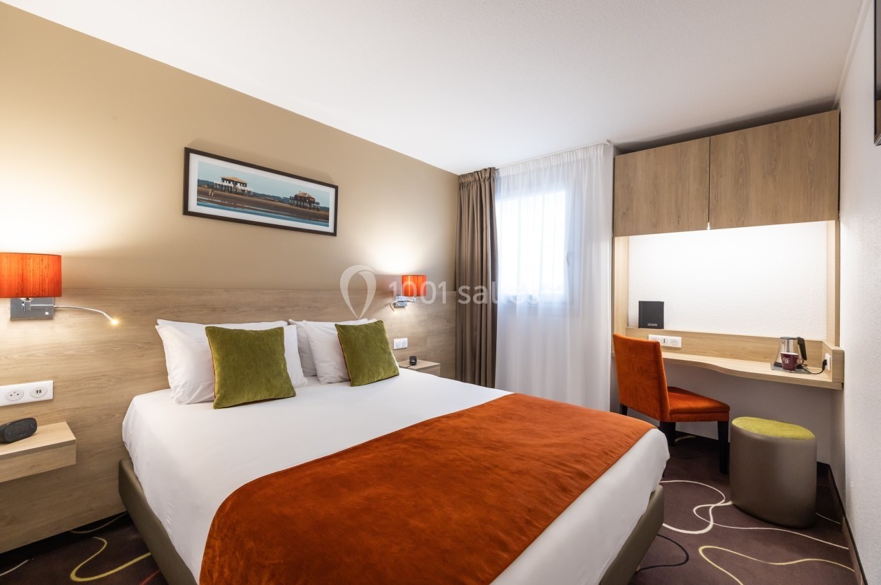 Chambre d'hôtel avec lit double, coussins colorés, bureau, chaise orange et éclairage chaleureux.