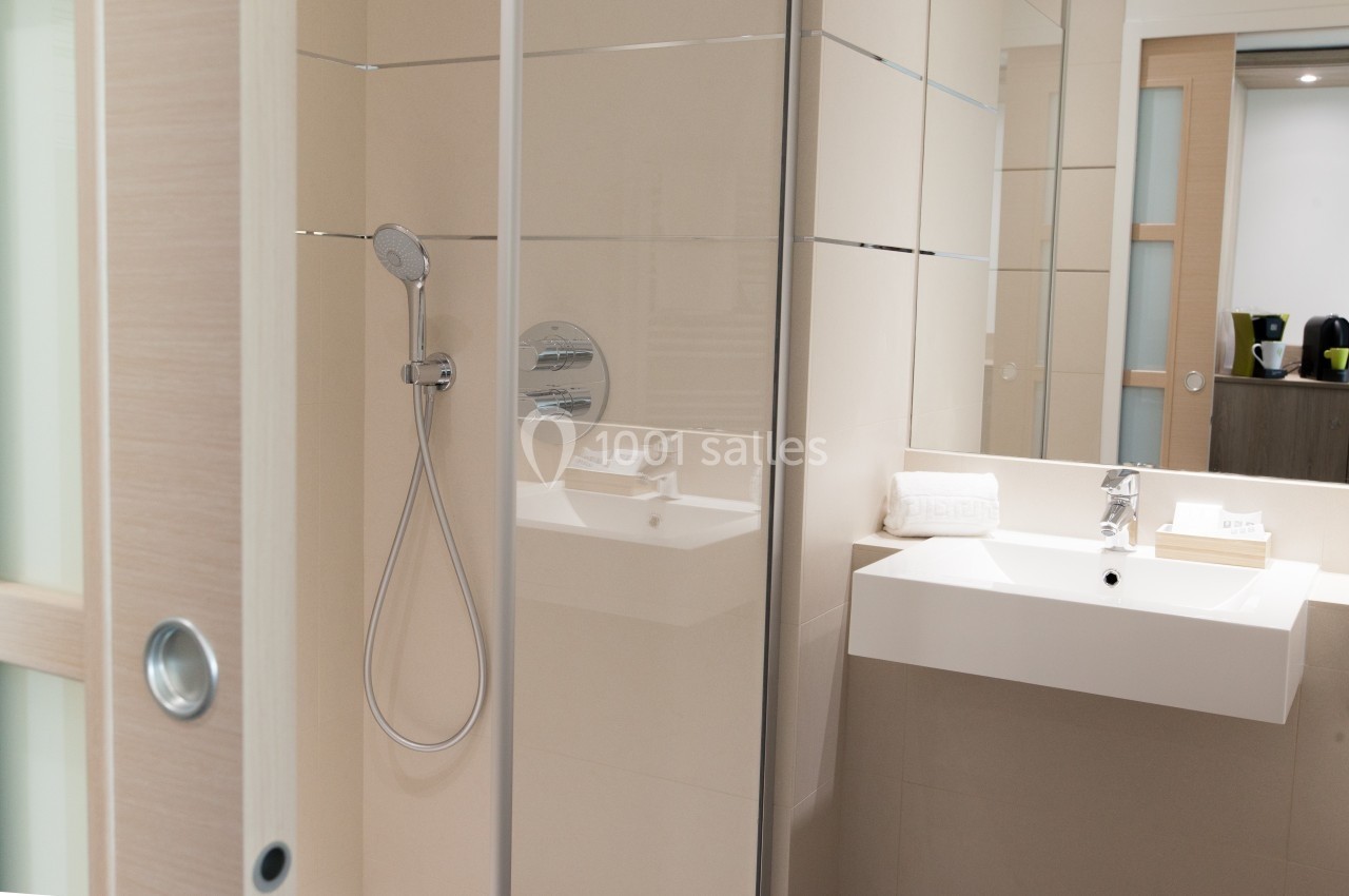 Douche moderne avec paroi vitrée, lavabo blanc et miroir dans une salle de bain lumineuse aux tons beiges.