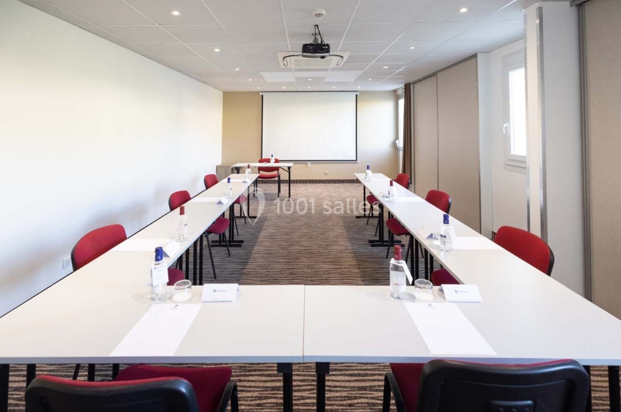 Salle de réunion lumineuse avec tables disposées en U, chaises rouges et écran de projection au fond.