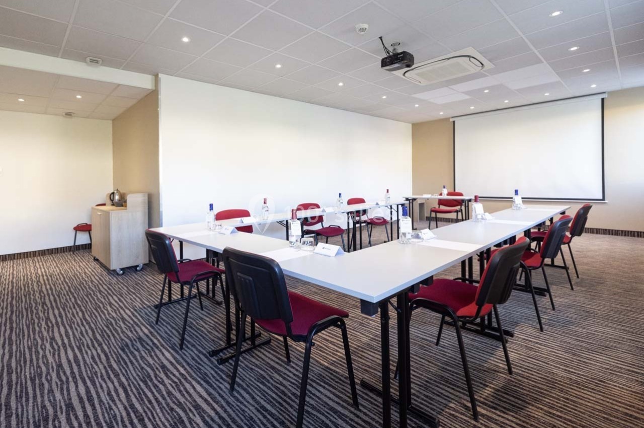Salle de réunion lumineuse avec tables disposées en U, chaises rouges, projecteur et écran de projection.