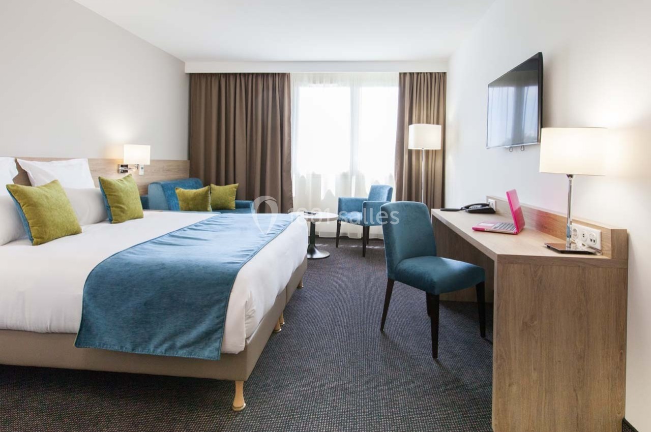 Chambre d'hôtel lumineuse avec lit double, bureau, fauteuils, télévision murale et grandes fenêtres avec rideaux.