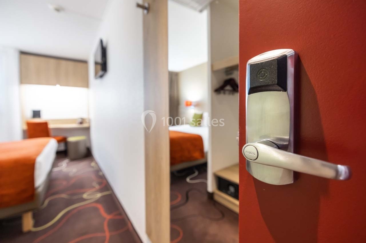 Vue d'une chambre d'hôtel avec porte entrouverte, lit double, bureau et décoration moderne.