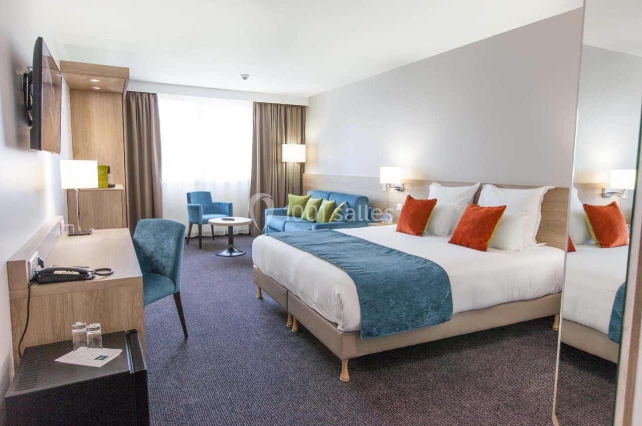 Chambre d'hôtel lumineuse avec lit double, canapé, bureau, télévision et grande fenêtre avec rideaux.