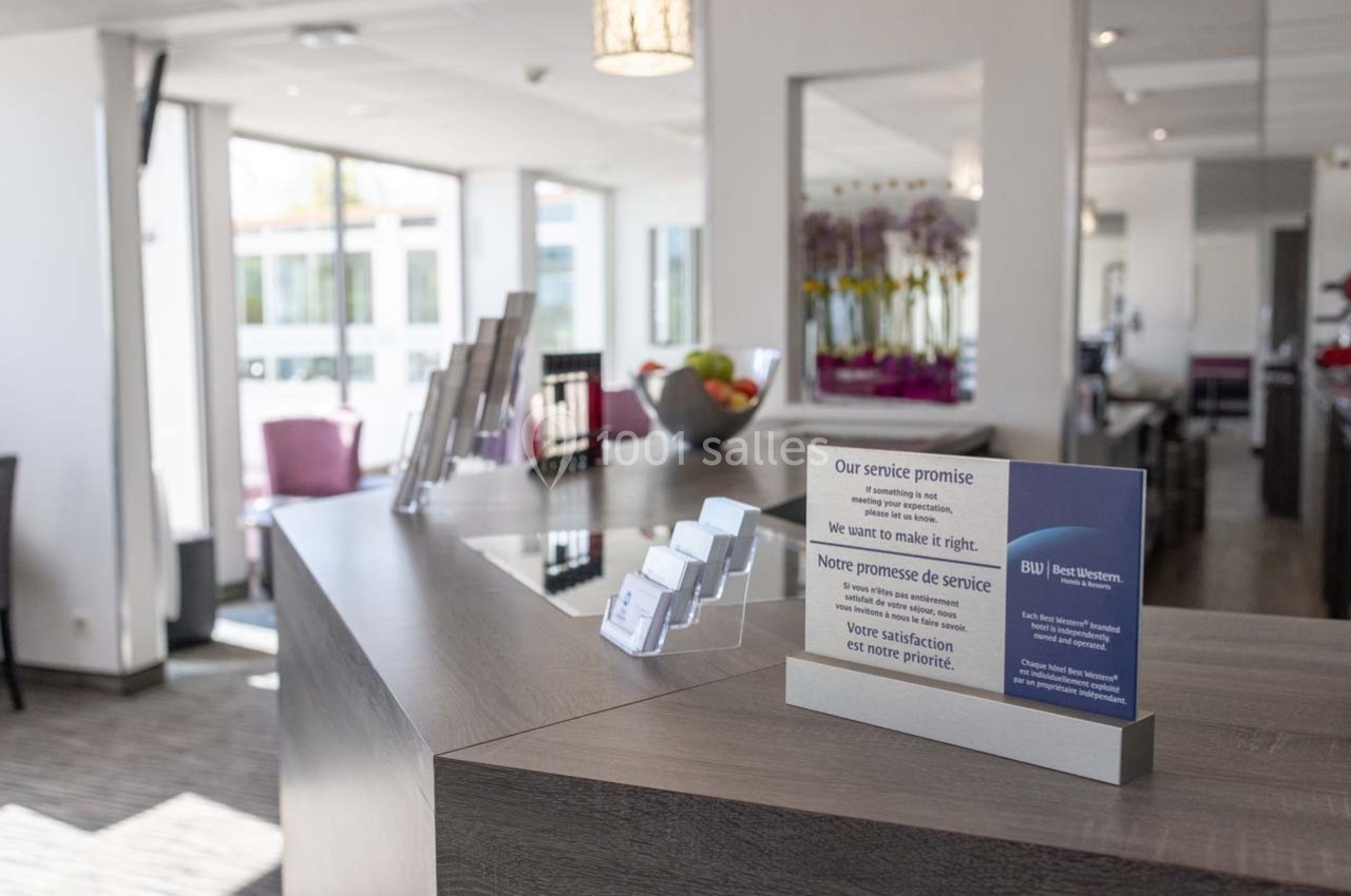 Réception d'hôtel avec un comptoir en bois, présentoirs de cartes de visite et un panneau affichant une promesse de service.