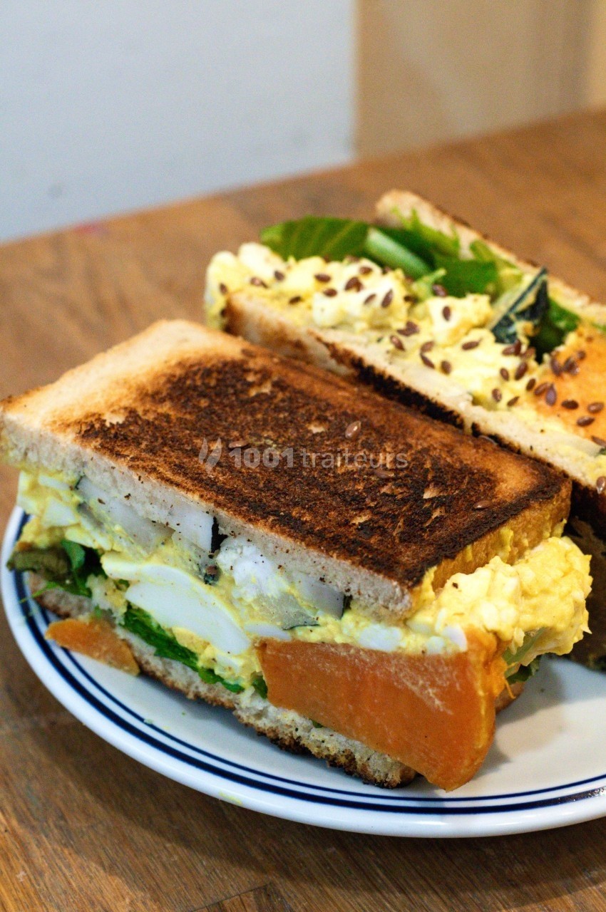 Sandwich garni d'œufs, légumes verts et graines, présenté sur une assiette blanche avec une tranche grillée.