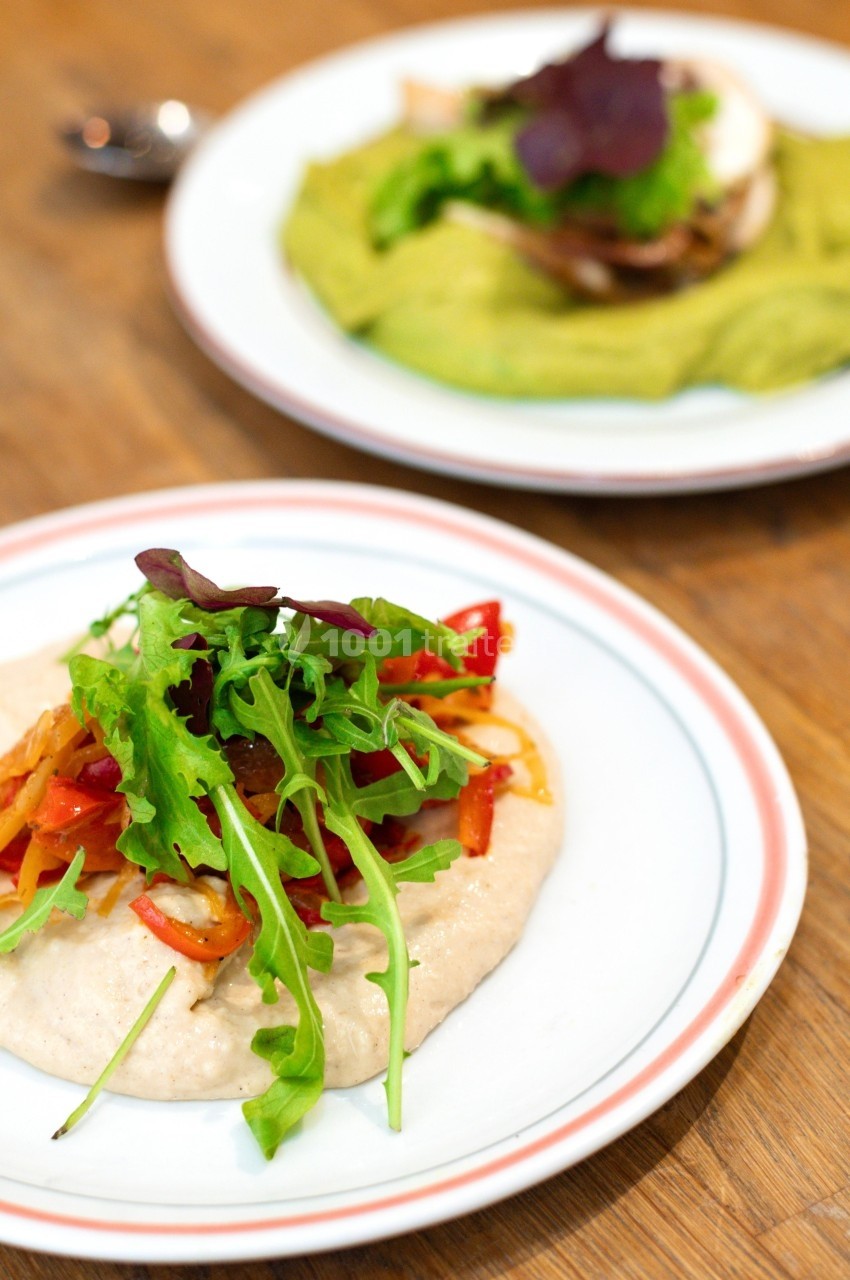 Assiette de purée garnie de légumes grillés et de jeunes pousses, avec un autre plat en arrière-plan flou.