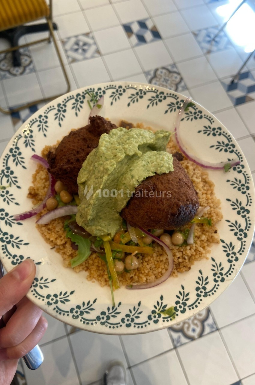 Assiette de couscous garnie de falafels, légumes, pois chiches et sauce verte, servie sur un carrelage blanc décoré.