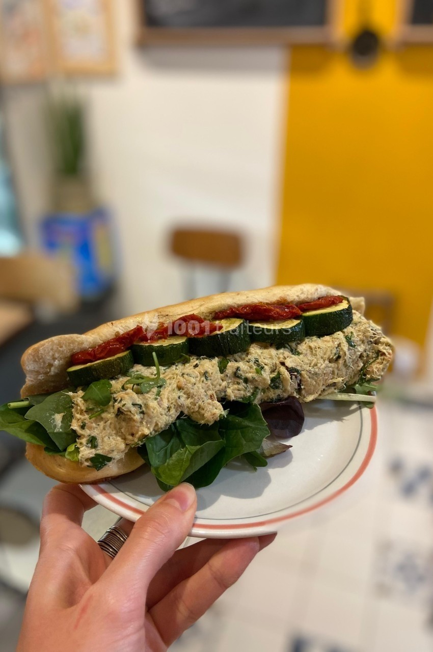 Sandwich garni de légumes grillés, feuilles de salade et tartinade, tenu sur une assiette dans un café lumineux.