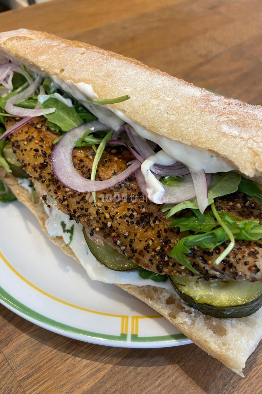 Sandwich garni de poisson grillé, rondelles d'oignon rouge, cornichons, roquette et sauce, servi sur une assiette.