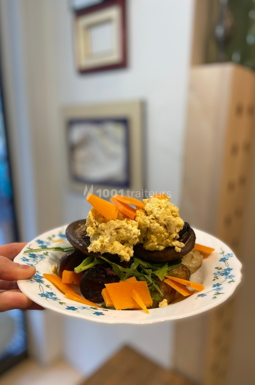 Assiette garnie de légumes grillés, roquette, morceaux de fromage et une préparation crémeuse, tenue devant un mur décoré.