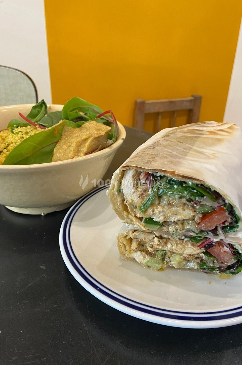 Un wrap garni de légumes et sauce, accompagné d'un bol de salade et houmous sur une table noire.