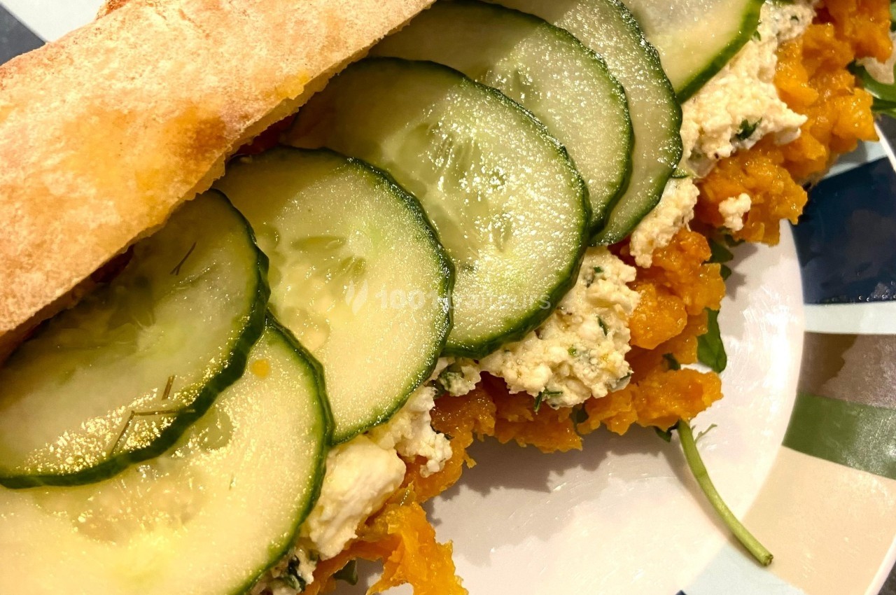 Sandwich garni de tranches de concombre, purée de légumes orange, fromage frais et roquette sur une assiette colorée.