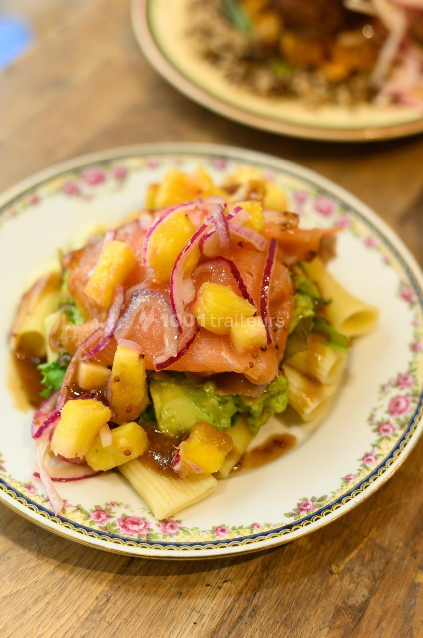 Assiette de pâtes garnie de saumon fumé, avocat, ananas, oignons rouges et sauce sur une table en bois.