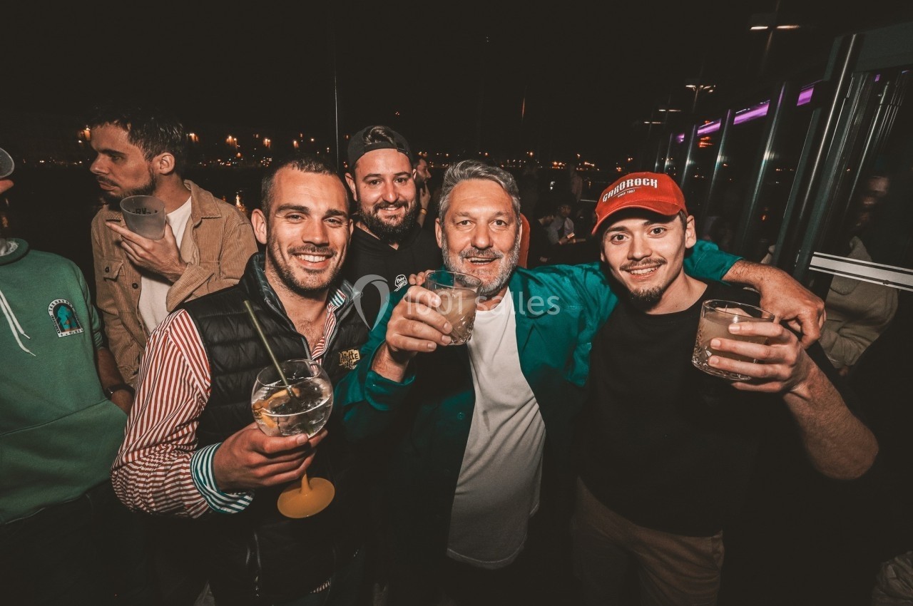 Quatre hommes souriants tenant des verres, réunis lors d'une soirée en extérieur avec des lumières en arrière-plan.