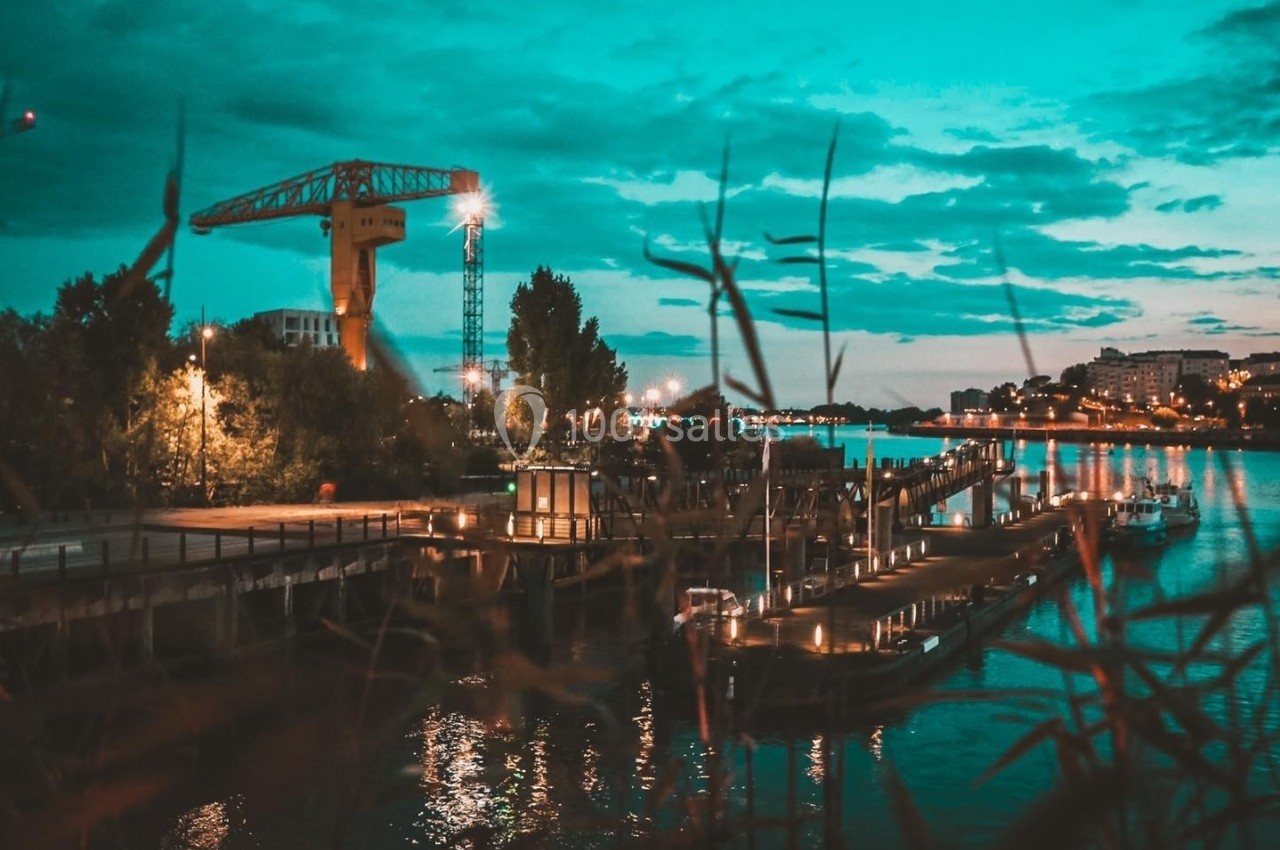Vue nocturne d'un port avec une grue illuminée, des quais éclairés et un ciel bleu-vert nuageux.