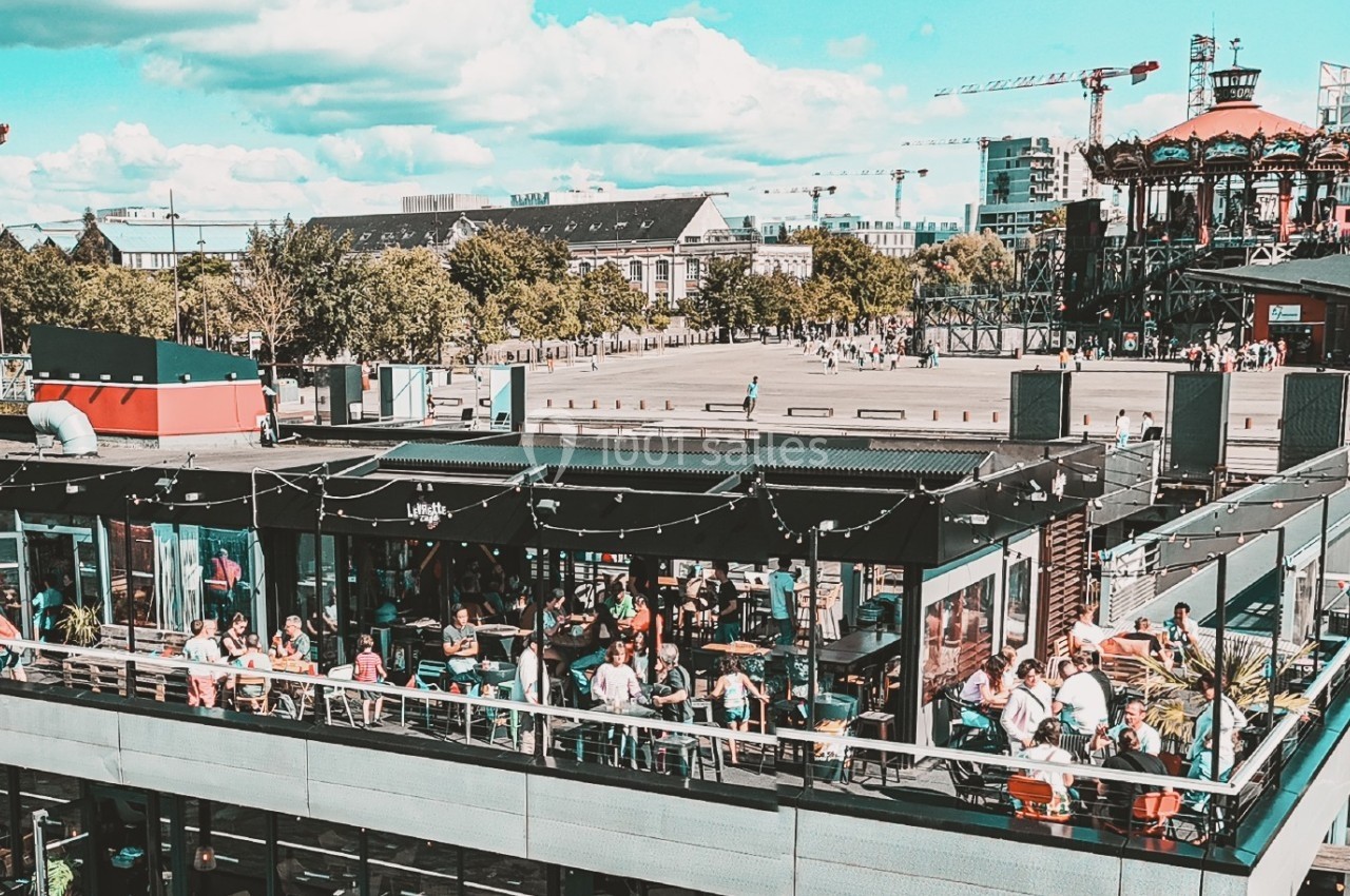 Terrasse animée d'un restaurant avec des clients, vue sur un espace urbain et des bâtiments en arrière-plan.