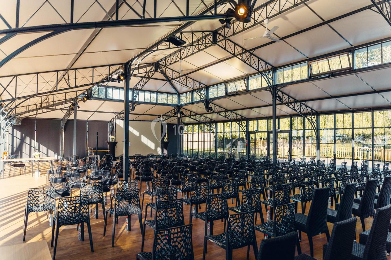 Salle lumineuse avec toit en verrière, rangées de chaises noires disposées face à une scène en bois.