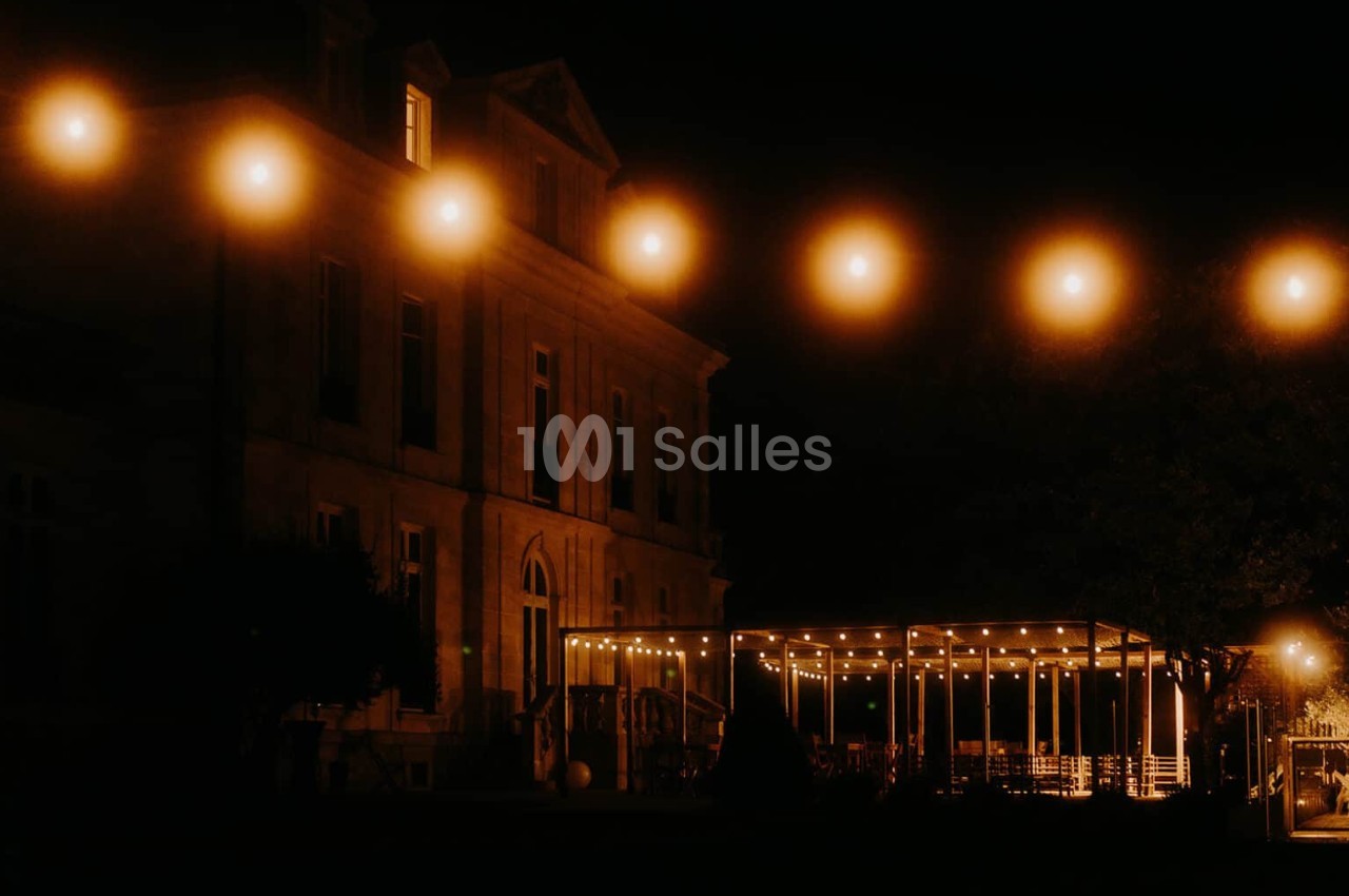 Façade d'un bâtiment ancien éclairé la nuit, avec une terrasse illuminée par des guirlandes lumineuses.